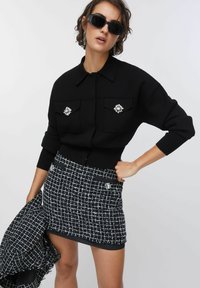 Giacca nera lavorata a maglia con tasche impreziosite da gioielli, abbinata a una minigonna in tweed a quadri bianco e neri. Tessuto liscio e strutturato.