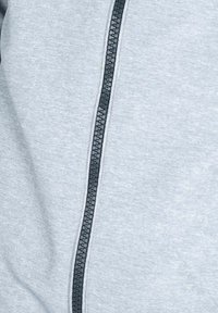 Lysegrå zip-up jakke med en sort lynlås og tekstureret stof. Designet inkluderer en glat overflade med subtile syninger.