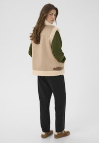 Beige vest met een fleecemanchet en zoom, gedragen over een groen longsleeve shirt. Zwarte broek en een paar bruine schoenen maken de outfit compleet.