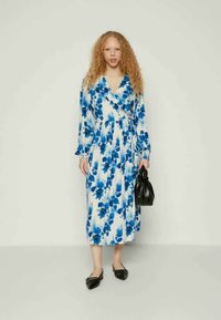 Φλοράλ φόρεμα τύπου wrapping σε λευκό και μπλε, μέχρι το γόνατο, με μακρύ μανίκι, από μαλακό υλικό. Το μοντέλο κρατά μια μαύρη τσάντα, συνδυασμένη με μαύρες φλατ παπούτσια.