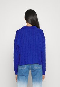 Blå kabelstickad tröja med en kortad design och en ribbad krage. Baksidan har ett texturerat mönster och avslappnade ärmar, tillsammans med ljusblåa jeans.