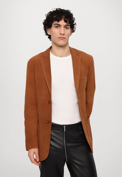 Brown suede blazer with two buttons, notch lapels, and a chest pocket, worn over a white textured shirt and black leather pants.