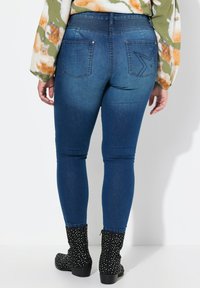 Jeans de mezclilla ajustados en un tono oscuro con una textura suave, que cuentan con bolsillos traseros y costuras decorativas. Combinados con botas de tobillo negras texturizadas.