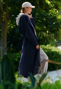 Jeune personne portant une casquette blanche et un long manteau sombre, parlant au téléphone en marchant à l'extérieur parmi des arbres et des plantes verts.