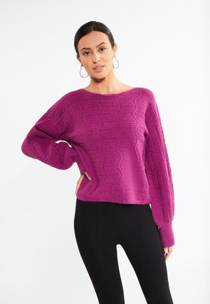 Frau mit dunklen Haaren und Creolen, die einen strukturierten lila Pullover und schwarze Hose trägt, posiert mit einer Hand auf der Hüfte.