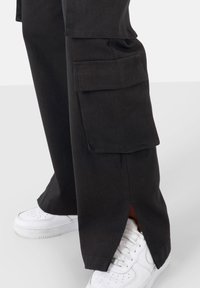 Zwarte cargobroek van duurzaam materiaal, met zijzakken en een gespleten zoom. Draagt met witte sneakers voor contrast.