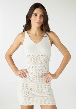 Donna con lunghi capelli scuri che indossa un abito bianco senza maniche in crochet con scollo a V, posando con le mani sui fianchi contro uno sfondo bianco uniforme.