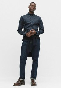 Man med mörkblå mönstrad skjorta med knappar, mörka jeans, bruna läderskor och en marinblå tröja knuten runt midjan, står avslappnat.