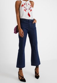 Weißes ärmelloses Top mit floralem Stickereimuster und Quasten, kombiniert mit hoch geschnittenen dunklen Jeans im Schlagstil und schwarzen Riemchenheels.