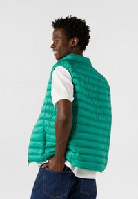 Chaleco acolchado color verde azulado con diseño acolchado de costuras horizontales, bolsillos con cremallera y amplios orificios para los brazos, superpuesto sobre una camiseta blanca.