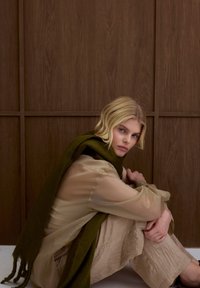 Blouse beige translucide avec une écharpe vert foncé, pantalon beige texturé, et un fond en bois minimaliste. Couleurs douces et matériaux naturels.