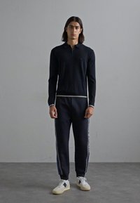 Un tânăr stă cu spatele la un zid simplu, purtând un top cu guler zip negru cu mânecă lungă, pantaloni de trening negri asortați cu dungi pe laterale și adidași albi.