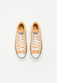 Geltoni kanvos sportbačiai su baltais raišteliais, juodu guminio pado ir baltais pirštų apsaugos. Ant liežuvėlio yra „Converse All Star“ logotipas.