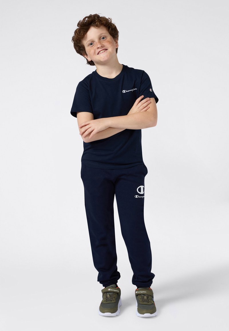 Glunderende jongen met krullend rood haar en sproeten staat met zijn armen gekruist, gekleed in een marineblauw Champion t-shirt, joggingsbroek en olijfgroene sneakers.