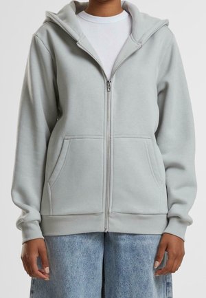 Personne portant un sweat à capuche gris clair zippé avec des poches devant, sur une chemise blanche et un jean bleu clair, montrée des épaules aux hanches.