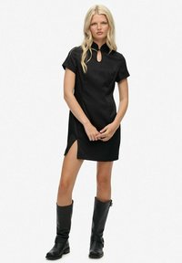 Schwarzes Kurzarmkleid mit hohem Kragen und Schlüsselaugen-Ausschnitt. Verfügt über einen Seitenschlitz und eine glatte Textur. Getragen mit kniehohen schwarzen Lederstiefeln.