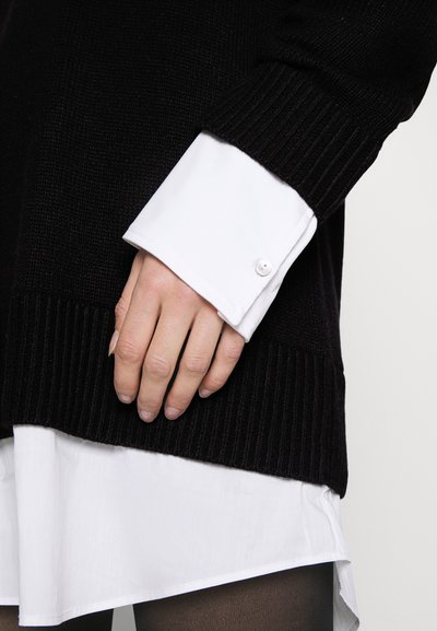 Schwarzer Strickpullover mit gerippten Bündchen über einem weißen Hemd mit Knöpfen. Die Schichten erzeugen einen Kontrast zwischen glatten und strukturierten Stoffen.
