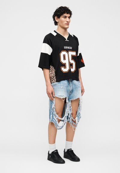 Άνδρας που φοράει μαύρο αθλητικό πουκάμισο με αριθμό 95 και πλευρές με τύπωμα λεοπάρδαλης, σε συνδυασμό με πολύ σκισμένα ανοιχτόχρωμα μπλε τζιν σορτς και μαύρα αθλητικά παπούτσια.