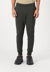 Mørkegrønne sweatpants med elastisk talje, tætsiddende pasform og manchetter ved anklerne, parret med sorte, mønstrede træningssko.