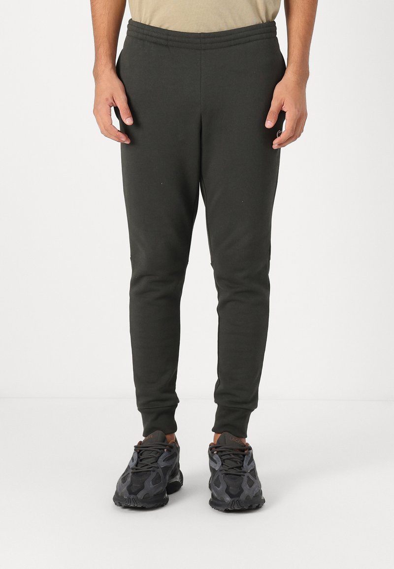 Mørkegrønne sweatpants med elastisk talje, tætsiddende pasform og manchetter ved anklerne, parret med sorte, mønstrede træningssko.