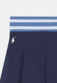 Laivaston sininen laskostettu hame, jossa on ribbahedelmäisen vaaleansinisen ja valkoisen raidallisen vyötärönauhan. Hameessa on myös pieni valkoinen logojen brodeeraus sivussa.