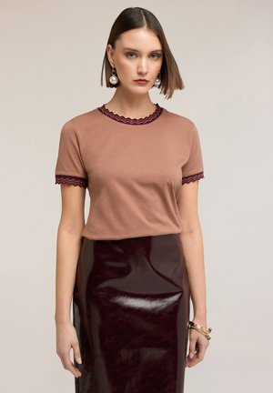 Kobieta w brązowym topie z krótkim rękawem z falistymi krawędziami i błyszczącą ciemną bordową skórzaną spódnicą, uzupełniona o perłowe kolczyki i bransoletki.