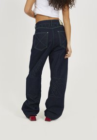 Dark blue denim jeans with green stitching, wide-leg design, two back pockets with x-stitch pattern, and a logo detail on the left leg.