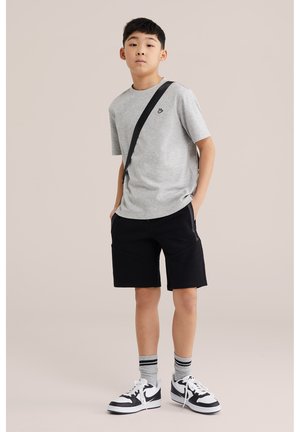 Ragazzo in piedi con le mani nelle tasche, indossa una maglietta grigia, pantaloncini neri, calzini grigi a righe, scarpe da ginnastica bianche e nere e una tracolla nera.