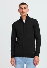 Svart zip-up genser med høy krage, ribbestrikkede mansjetter og kant, laget av et mykt materiale med en glatt tekstur.