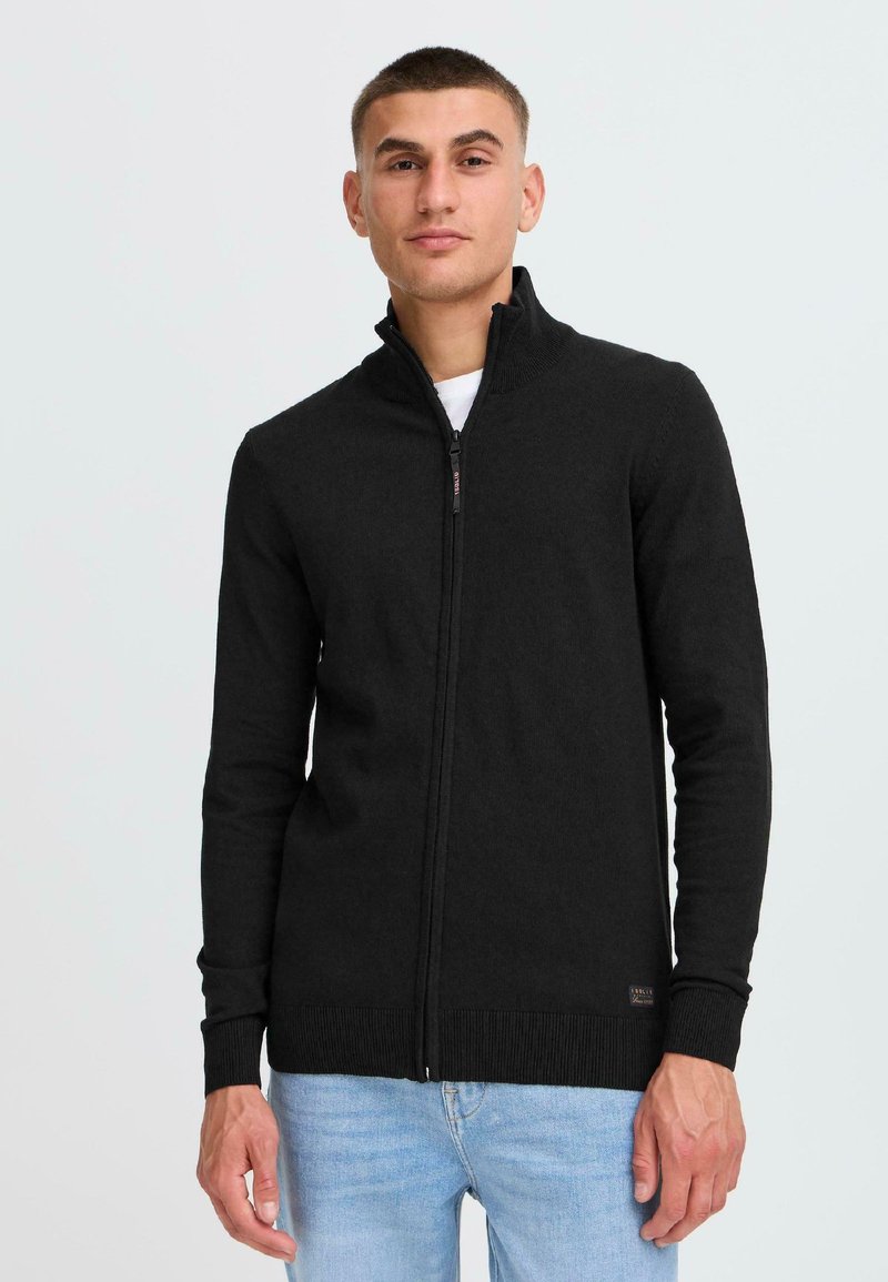 Svart zip-up genser med høy krage, ribbestrikkede mansjetter og kant, laget av et mykt materiale med en glatt tekstur.