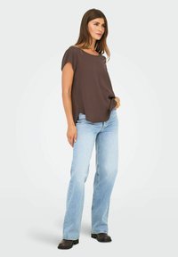 Donna che indossa un top corto marrone largo con maniche corte, jeans dritti azzurri chiari e stivali alla caviglia marrone scuro, in piedi con una mano in tasca.