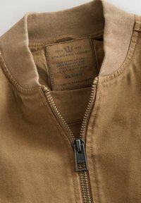 Tan bomberjack met een gladde stofafwerking, zichtbare rits en geribbelde kraag. Het binnenlabel zegt "STANDARD ISSUE" in het blauw, maat medium.