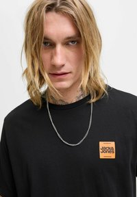Schwarzes T-Shirt mit einem rechteckigen orangenen Logo auf der linken Brustseite. Es hat einen runden Ausschnitt und einen weichen Stoff. Eine silberne Kettenkette wird getragen.