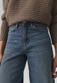 Denim jeans with a high waist, featuring a light blue wash, five pockets, and contrast stitching. Worn with a brown ribbed knit sweater.