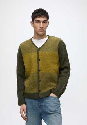 Jeune homme portant un cardigan vert et jaune en laine douce boutonné par-dessus une chemise blanche et un jean bleu, debout avec une main dans la poche.