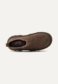 Scarpa slip-on in suede marrone con punta rotonda, pannelli elastici laterali e suola in gomma testurizzata con un lieve tacco. Logo UGG visibile all'interno.