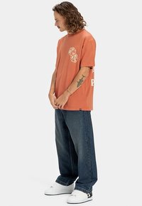 Mand med skulderlang krøllet hår, iført en løs orange t-shirt, brede mørke jeans, hvide sneakers og en haj-tatovering på sin venstre underarm.