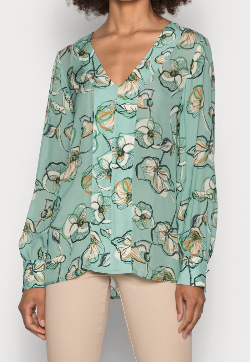 Mujer que lleva una blusa floral de color teal claro, de mangas largas y cuello en V, combinada con pantalones beiges, frente a un fondo claro y liso.