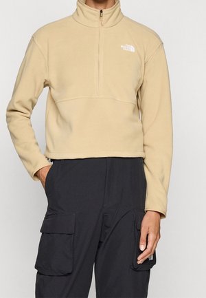 Osoba ubrana w beżowy polar z krótkim suwakiem i czarne spodnie cargo, z rękami w kieszeniach, na jednolitym tle.