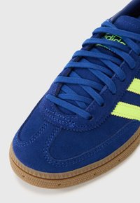 Sneaker azul de camurça com riscas amarelas, parte superior texturizada, biqueira redonda, atacadores planos e sola de borracha. Vista aproximada mostrando os detalhes da costura.