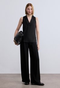 Μαύρο αμάνικο τοπ με V-λαιμόκοψη, συνδυασμένο με μαύρο παντελόνι φαρδιάς γραμμής και μαύρο τσαντάκι χειρός. Λείο ύφασμα, μοντέρνο κόψιμο, μινιμαλιστικός σχεδιασμός.