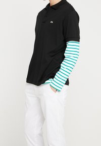 Chemise polo noire avec un logo vert et une chemise à manches longues blanche en dessous, dotée de rayures turquoise et blanches ; portée avec un pantalon blanc.