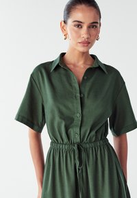 Zielona sukienka zapinana na guziki z krótkimi rękawami, regulowanym paskiem w talii i miękką teksturą. Posiada kołnierzyk i prosty zamek na guziki.