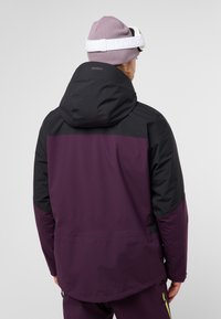 Giacca isolante viola e nera con cappuccio, caratterizzata da una consistenza liscia, orlo regolabile e blocchi di colore a contrasto.