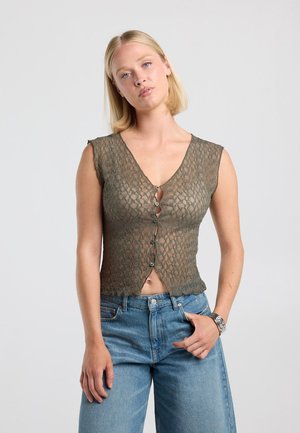 Femme aux cheveux blonds portant un haut transparent, sans manches, à motifs et boutonné, ainsi qu'un jean bleu, debout devant un fond uni clair.