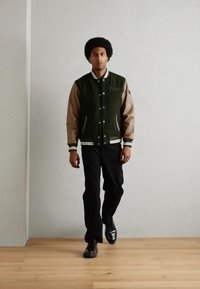 Chaqueta varsity para hombre con un cuerpo de lana verde oscuro y mangas de cuero marrón, con rayas blancas en los puños y el cuello. Pantalones y zapatos negros.