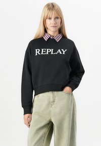 Zwarte cropped sweatshirt met het "REPLAY" logo in het wit, gecombineerd met een roze en witte geruite blouse met kraag, en lichtgroene wijde jeans.