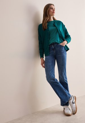 Teal cardigan over en matchende top, parret med lyseblå jeans og hvide sneakers. Glat tekstur med knapdetaljer på cardiganen.