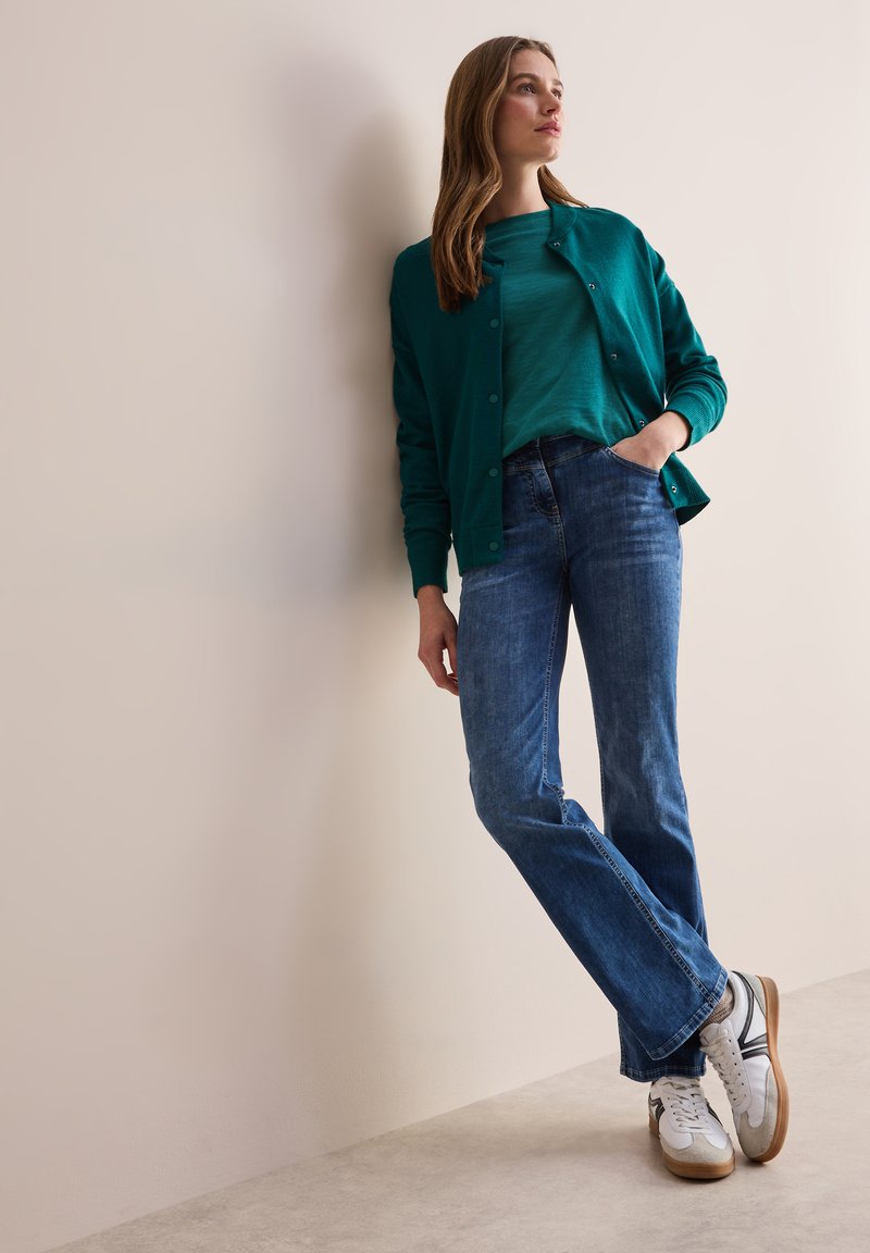 Sinitrans cardiganin päällä on yhteensopiva toppi, yhdistettynä vaaleansinisiiin farkkuihin ja valkoisiin lenkkareihin. Cardiganissa on sileä pinta ja nappidetaljit.