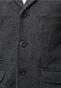 Blazer gris texturizado con cuello con muesca, que presenta tres botones negros, dos bolsillos frontales y un patrón tejido sutil.